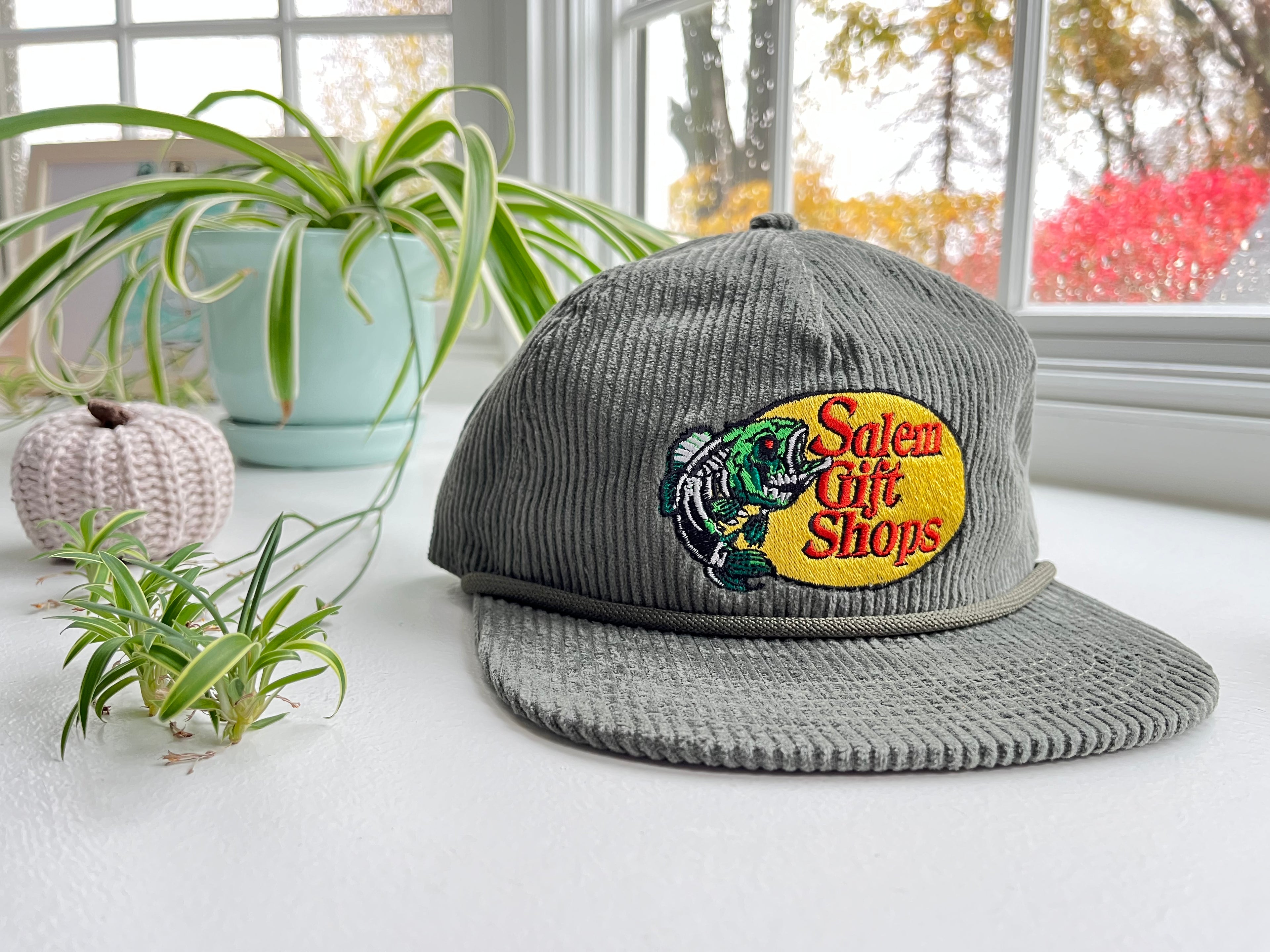Green corduroy cap with embroidered logo on a surface with plants and a window in the background