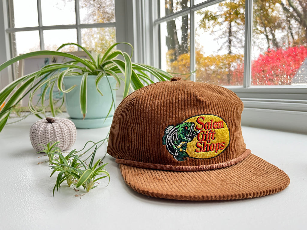 Brown corduroy cap with embroidered logo on a white surface with plants and a window in the background